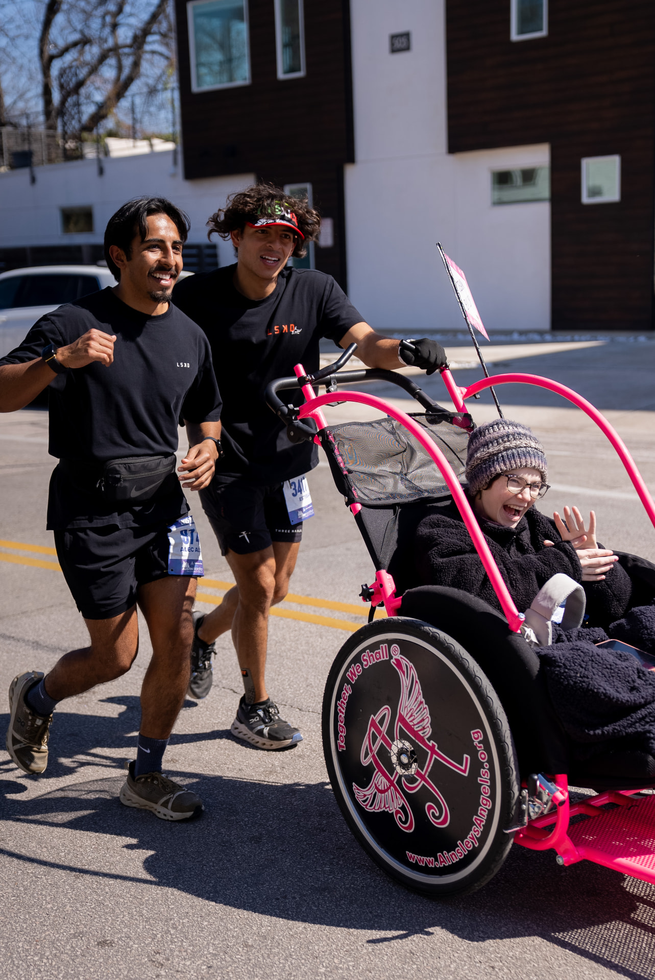 Two runners pushing an Ainsley's Angels athlete in a pink racing chair